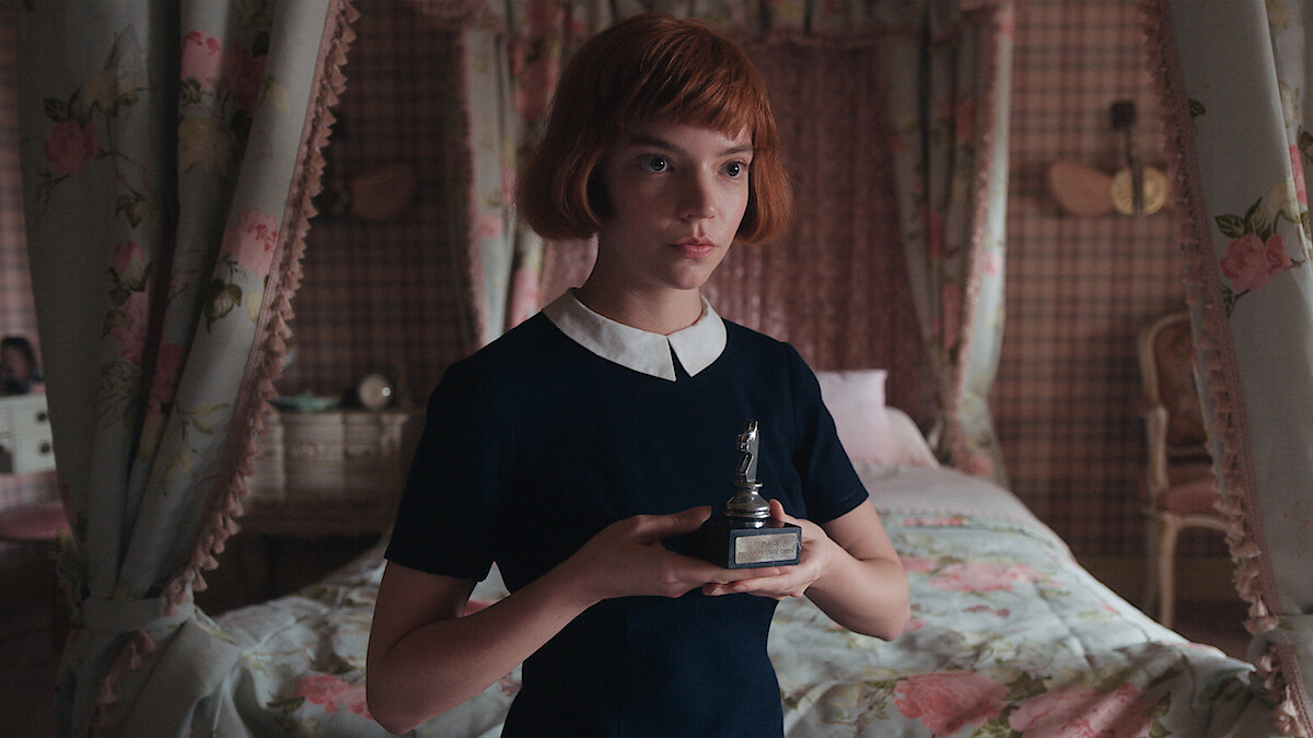 Young girl with short red hair in a dark dress holds a chess trophy in a vintage, floral-decorated bedroom with a canopied bed and patterned wallpaper.