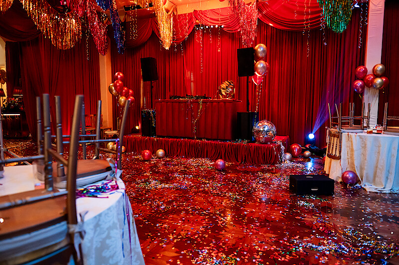 Um salão em tons de vermelho onde uma festa acabou de terminar tem papel picado e adereços brilhantes por toda parte