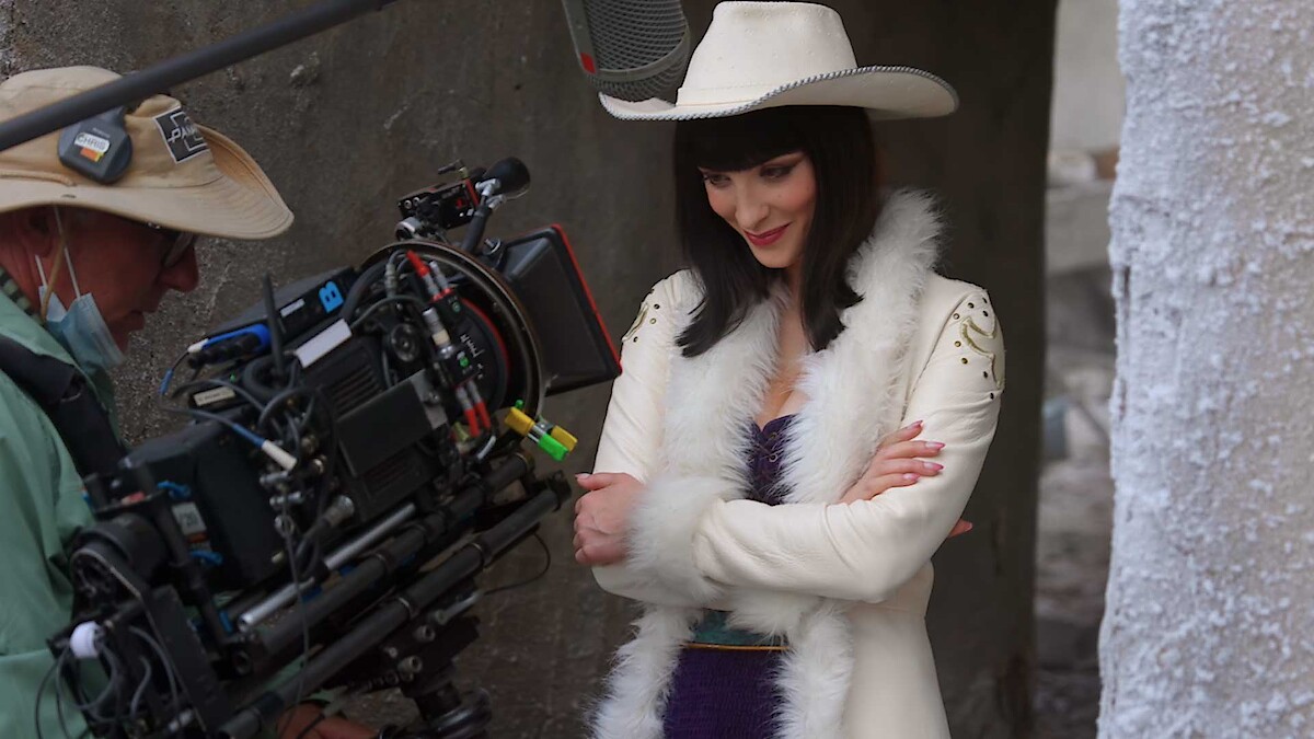 A woman in a white fur coat and cowboy hat poses on a film set, smiling with arms crossed, while a cameraman in outdoor gear films her in a narrow concrete corridor.