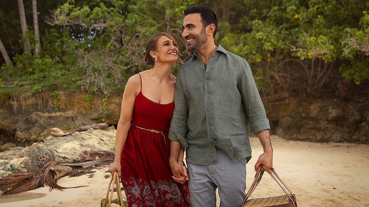 A happy couple holding hands and carrying beach bags, walking on a sandy beach with trees and rocks in the background, enjoying a pleasant outdoor day together.