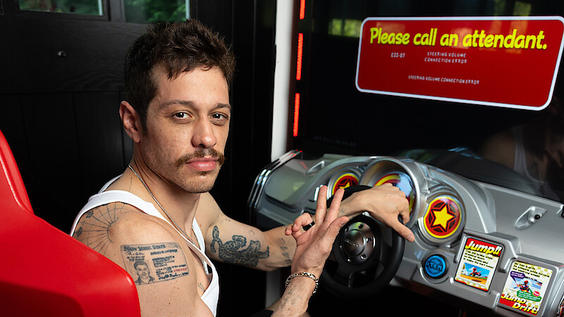 Man with tattoos in a white tank top sitting at an arcade driving game machine, steering with one hand and making a hand gesture, with a “Please call an attendant” message on the screen in a casual indoor setting.