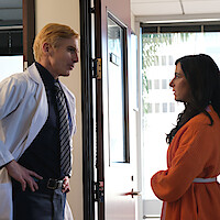 A doctor in a white coat talks to a woman in an orange sweater inside an office with large windows and a modern interior, both standing near an open door, appearing engaged in conversation.