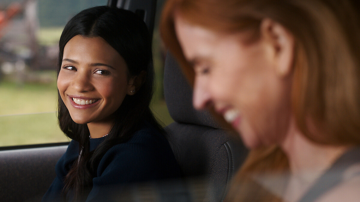 Two women sitting in a car, one with dark hair smiling warmly at the other, who has red hair and is laughing. They appear to be enjoying a friendly moment together while parked or driving during the daytime.