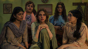 Five women sit closely together in a dimly lit room with green walls, looking serious and contemplative. They wear traditional clothing and appear to be engaged in an emotional conversation or moment.
