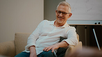 Smiling older man with glasses and white shirt sits relaxed in a beige armchair in a modern office or therapy room, with a flip chart in the background.