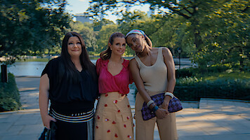 Three women standing close together outdoors on a sunny day in a lush park, smiling and posing for a photo with trees, greenery, and a body of water in the blurred background.