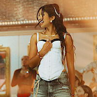 Woman with long hair in a white corset top and jeans, accessorized with gold jewelry, smiling in a lively indoor setting with people and art in the background, warm lighting.