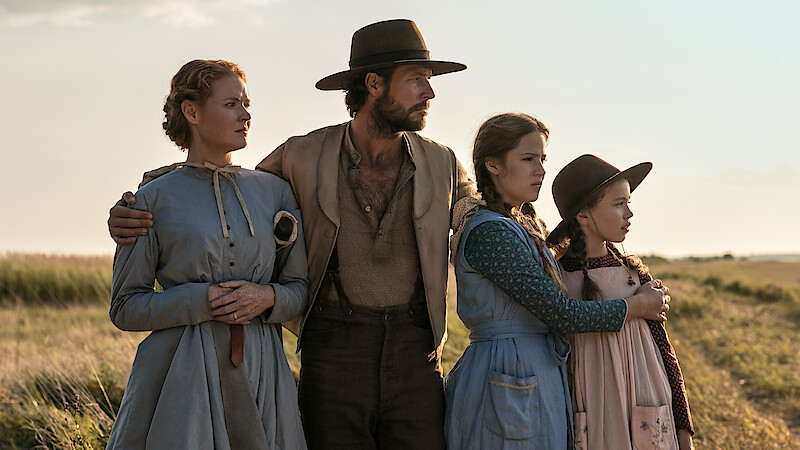 A man and three girls in old-fashioned clothing stand close together in a grassy field, looking intently into the distance under an open sky, evoking a historical or pioneer setting.