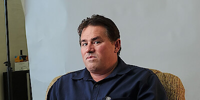 A man with brown hair in a blue collared shirt sits in front of a white background and looks directly to camera.