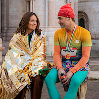 Two marathon runners sit on a stone bench, smiling at each other. One is wrapped in a shiny gold thermal blanket, the other is in bright running clothes with a medal and drink bottle.