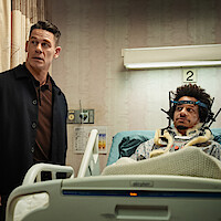A man wearing a dark jacket stands beside a hospital bed where another man lies with a head brace and neck support, surrounded by medical equipment in a hospital room, patient appears injured.