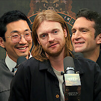 Lee Sung Jin, Finneas O'Connell, and Jake Schreier, smiling with microphones in front of them and a decorative backdrop behind.