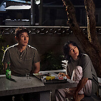 Two people sit at a stone table outdoors at night, looking up. The table holds a green bottle, a glass, snacks and food wrappers. A tree and concrete wall are in the background, suggesting a quiet, casual setting.