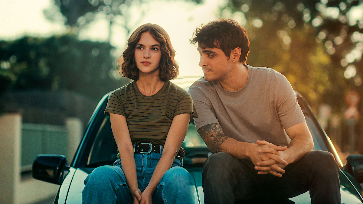 A young woman and man sit on the hood of a car outdoors, surrounded by trees and soft sunlight, appearing relaxed and engaged in conversation in a quiet suburban setting.