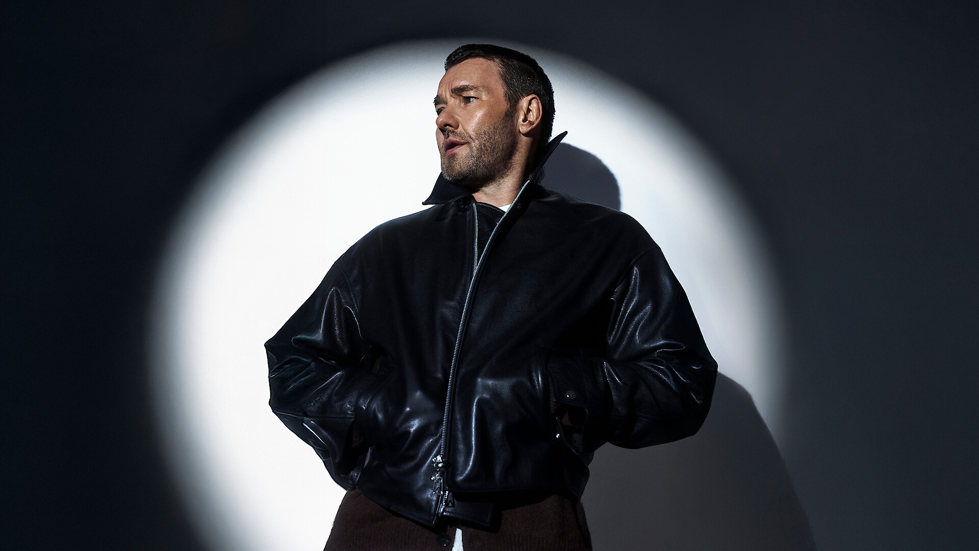 Joel Edgerton illuminated by a spotlight in front of a white wall wearing a black leather jacket looking to the side. 