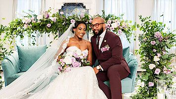 Bride and groom sitting on a light blue couch, both smiling, surrounded by lush green and purple floral arrangements in a bright and elegant indoor wedding setting.