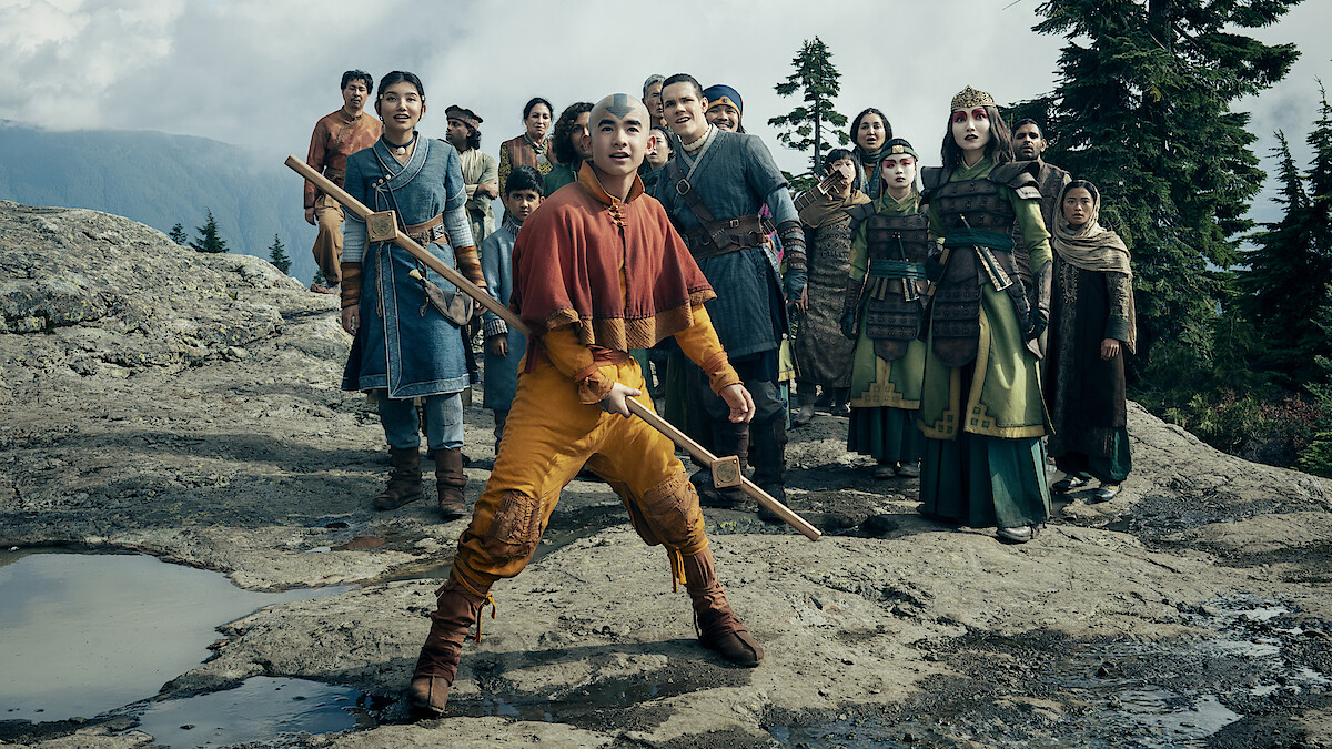 A group of people in costumes stand outdoors on rocky terrain with trees and mountains in the background, the central figure holding a staff and looking alert.