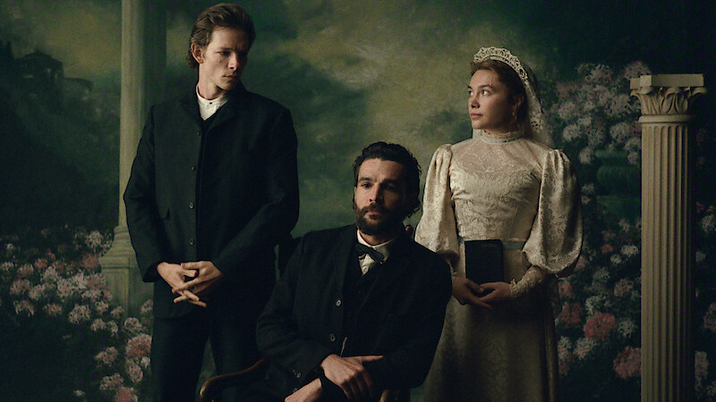 Three people in vintage clothing pose for a formal portrait with a scenic painted backdrop and decorative column, evoking a 19th-century setting. One woman in a lace dress and two men in dark suits.