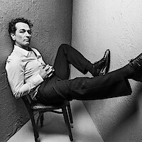 Man in formal attire sits on chair in a corner, legs extended and propped up against the walls. Black and white photo with dramatic lighting, creating a stylish, contemplative, slightly edgy mood in a minimal setting.