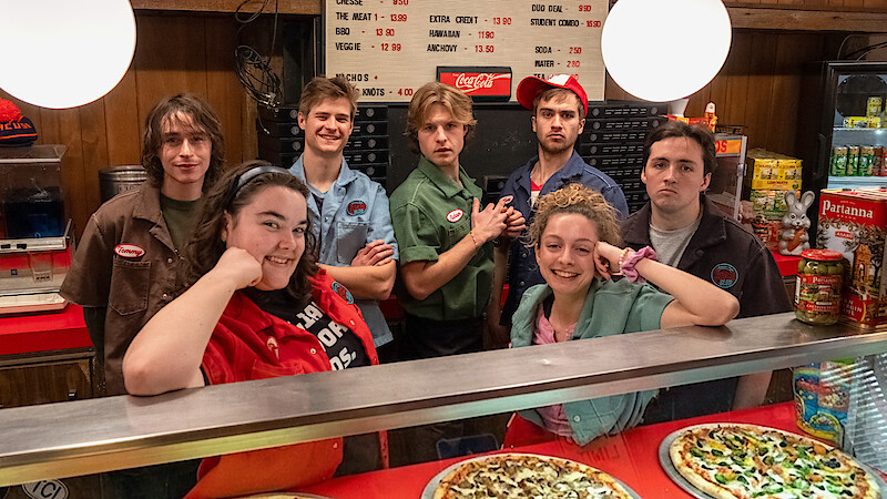Seven young people in pizza shop uniforms stand behind a glass counter with pizzas, smiling and posing for a photo; wood paneling, a menu board, and snack shelves are visible in the cozy, retro-styled restaurant environment.