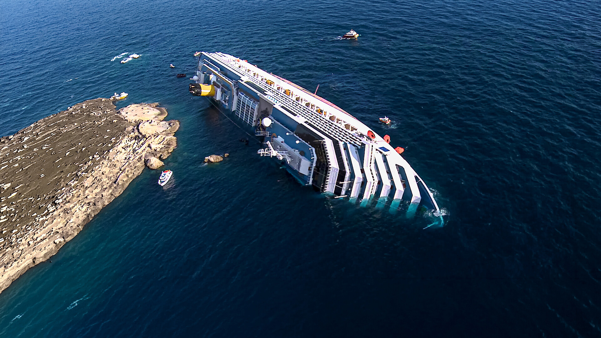 Large cruise ship partially submerged and tilted on its side near rocky shoreline in open ocean, surrounded by rescue boats and debris, with blue water and clear sky.