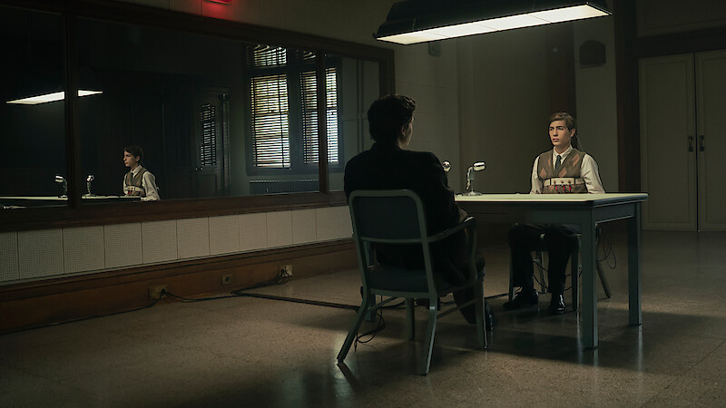 Two people sit across from each other at a table in a dimly lit interrogation room, with a large mirror on the wall reflecting the scene and a microphone on the table, creating a serious and tense atmosphere.