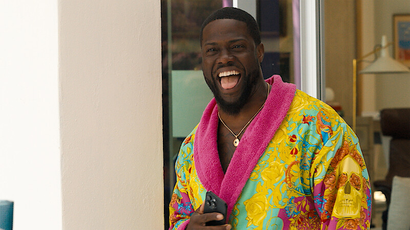 Man standing indoors near a doorway, laughing joyfully, wearing a colorful bathrobe with a bright pink collar and holding a smartphone, with modern home furnishings in the background.