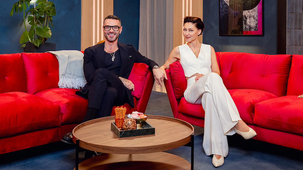 Two people sit on bright red sofas in a stylish living room with a round coffee table holding snacks, decorative items, and a plant in the background. The lighting is warm and the setting is modern and inviting.