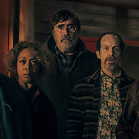 Three adults indoors stand closely together, looking ahead with surprised or concerned expressions; the environment suggests a dimly lit, rustic wooden room or cabin.