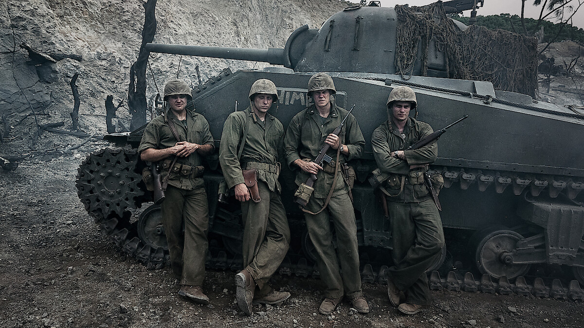 Four soldiers in WWII uniforms stand in front of a tank in a war-torn, smoky environment, suggesting a battlefield setting.