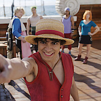Smiling young man in a straw hat and red vest poses on a wooden ship deck with four friends behind him, ocean visible in the background, all in colorful costumes, suggesting an adventurous nautical setting.