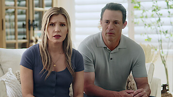 A concerned woman and man sit together on a sofa in a bright, modern living room with light walls, plants, and wooden furniture visible in the background.