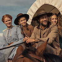 A pioneer family in old-fashioned clothing sits together on a covered wagon, smiling outdoors under a clear sky, suggesting a journey or migration in a rural historical setting.