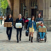 Five people carrying boxes and bags, including one in a wheelchair, walk together outside a historic stone building with arches and lanterns, suggesting a college or university move-in day.