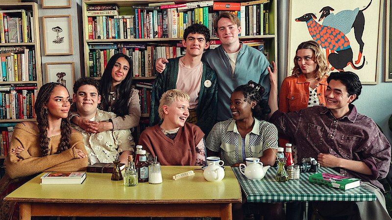 A diverse group of eight young people sitting and standing around a table in a cozy room with bookshelves and art, sharing a friendly moment, some smiling and talking, with tea and condiments on the table.