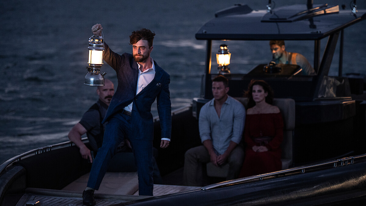 A man in a blue suit holds up a lantern on a small boat at dusk, surrounded by four other people sitting and watching, with a dark sea and another boat in the background.