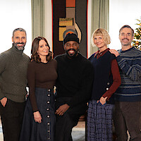 Five adults pose together and smile in a warmly lit room decorated for the holidays, with a Christmas tree in the background and elegant furnishings, suggesting a festive and friendly gathering.