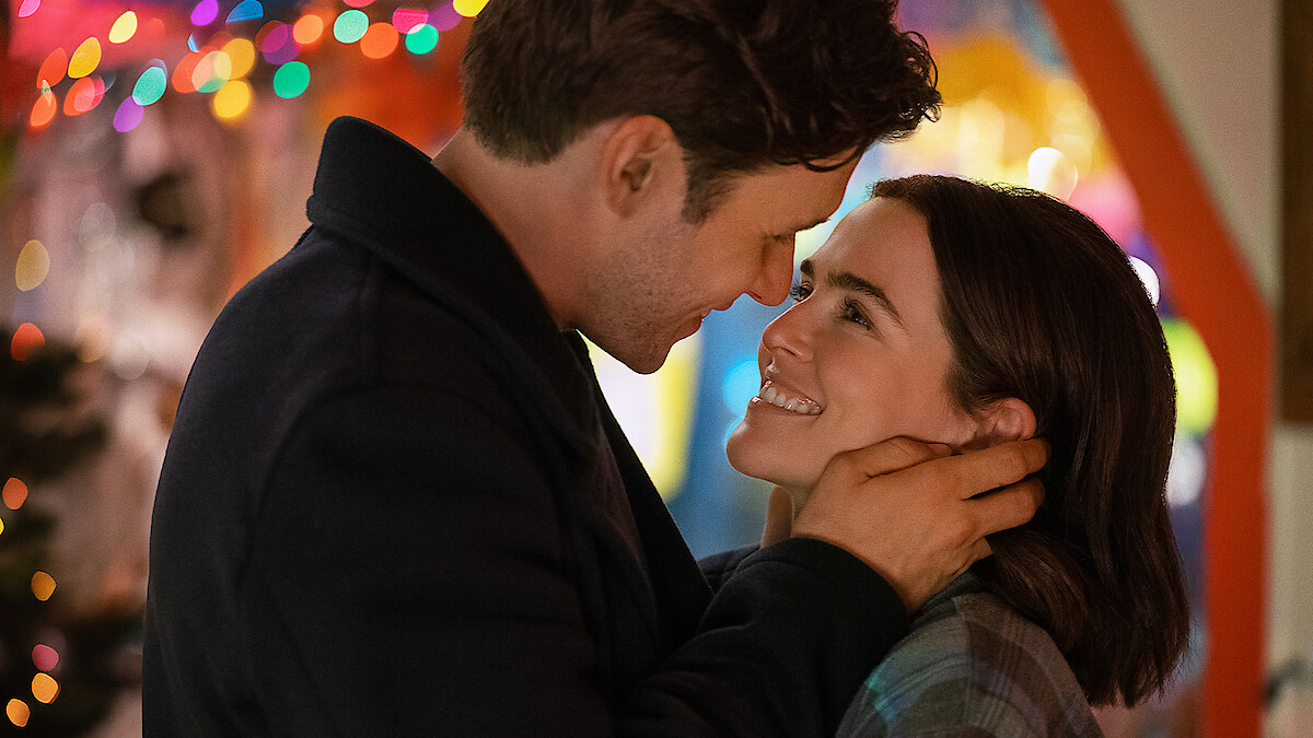 A couple embraces and smiles at each other lovingly indoors, surrounded by colorful holiday lights in a festive, warm setting.