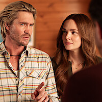 A man in a plaid shirt gestures while speaking, standing next to a woman with long hair. They are indoors in a wood-paneled room, and part of another person is visible in the foreground.