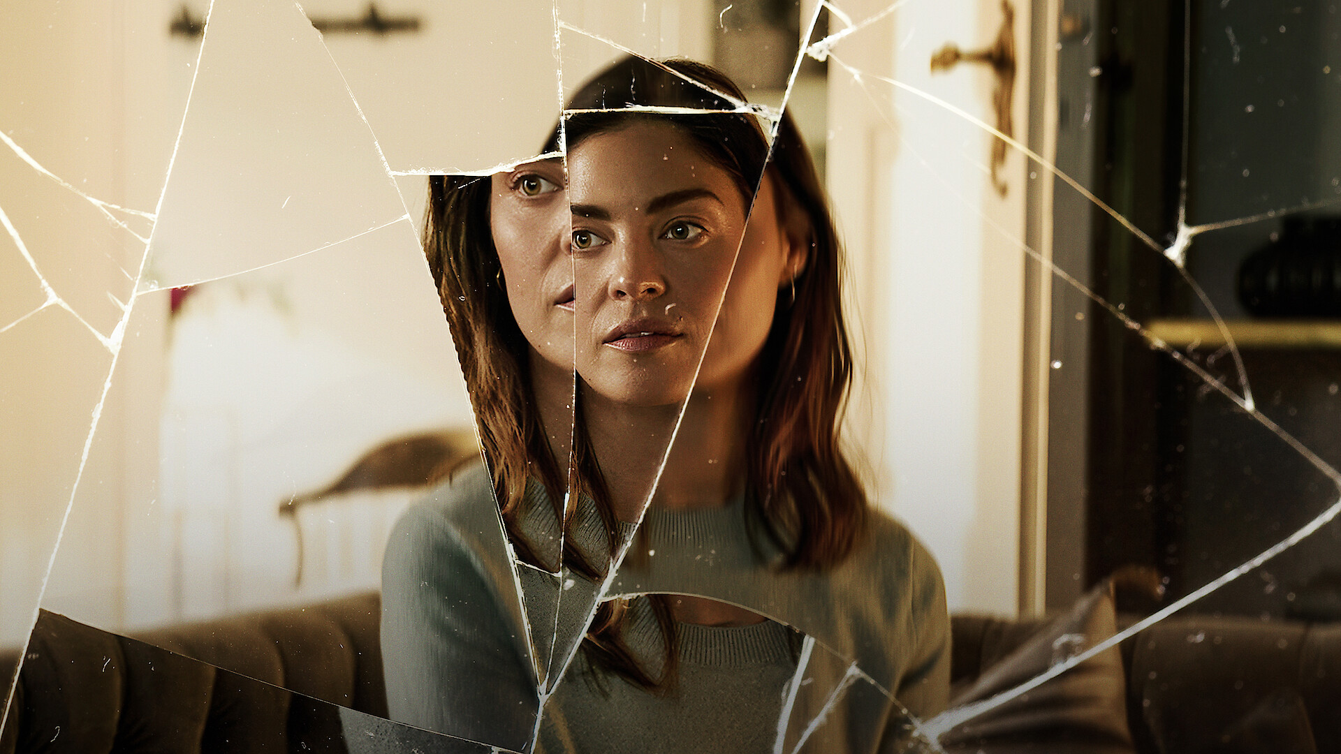 Woman sitting on a couch in a warmly lit room, her face reflected and fragmented in shards of a broken mirror, creating a surreal and disorienting visual effect.