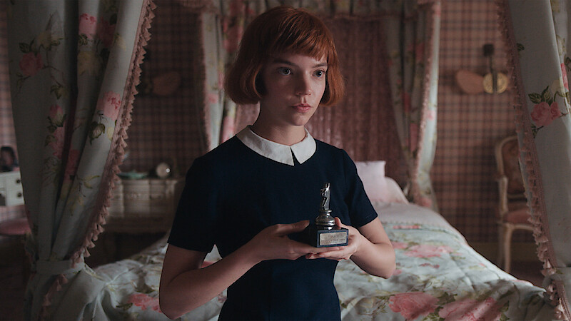 Young girl with short red hair in a dark dress holds a chess trophy in a vintage, floral-decorated bedroom with a canopied bed and patterned wallpaper.