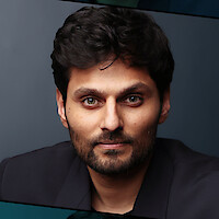 Man with dark hair and beard wearing a dark blazer, sitting indoors against a plain, dark background, looking directly at the camera with a neutral expression. The setting appears professional and studio-lit.