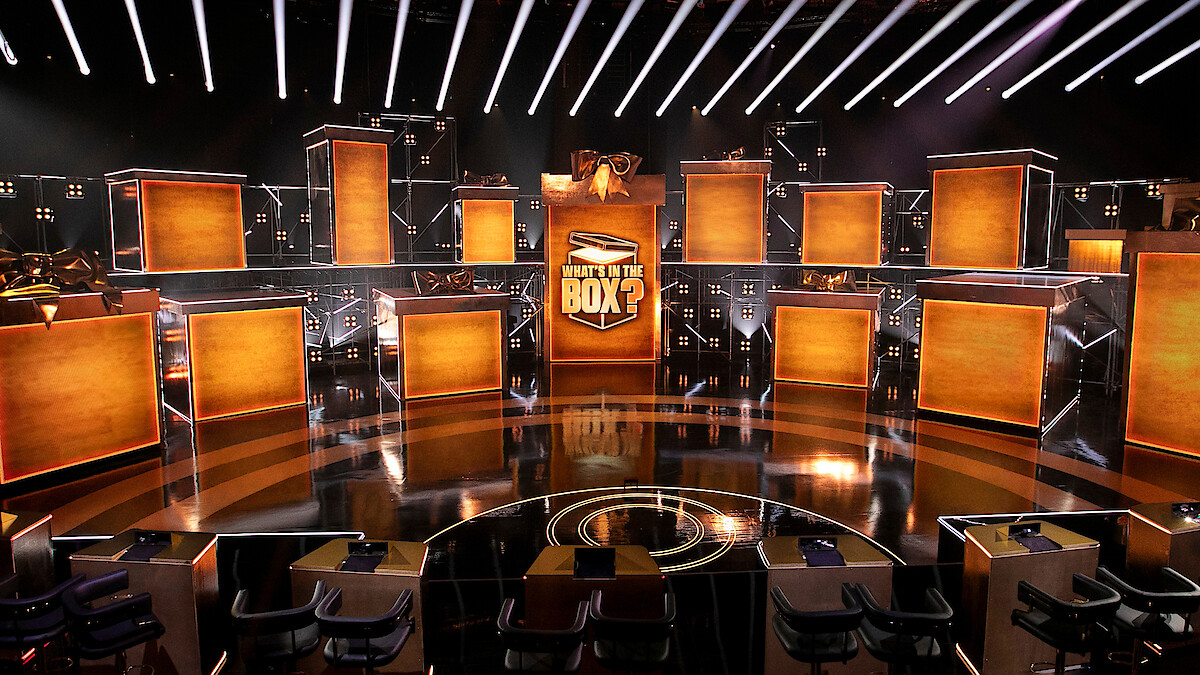 Game show stage set with multiple large illuminated boxes, dramatic lighting, and a central sign reading "What's in the Box?" Modern design, energetic atmosphere, empty seats and anticipation in the environment.