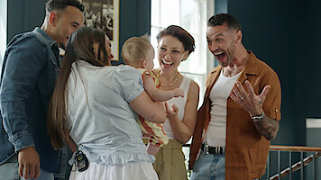 Four adults gathered indoors around a smiling baby, sharing a joyful moment in a brightly lit room with dark walls and framed pictures, one person expressing surprise and excitement.