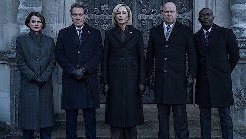 Three people in dark formal coats and gloves stand outside in front of an old stone building and ornate doors, posing seriously for a photograph in a somber or official setting.