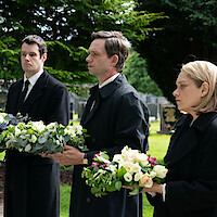 Three solemn people in black coats holding flower arrangements stand in a cemetery, surrounded by gravestones and green trees, attending a funeral on a cloudy day.