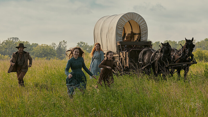 Four people in pioneer-era clothing run through a grassy field beside a covered wagon pulled by two horses, with trees and sky in the background, evoking a historical or frontier setting.
