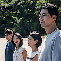 Four adults stand outdoors on a sunny day, with lush green trees in the background. They appear relaxed and are looking in various directions, suggesting a casual moment in nature.