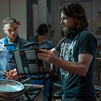 A director on set in casual dress shows an actor a tablet as they work together on a shot.