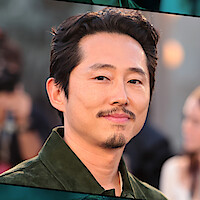 Steven Yeun with short dark hair and goatee wearing a green shirt, posing outdoors at an event with a blurred crowd and warm lighting in the background.